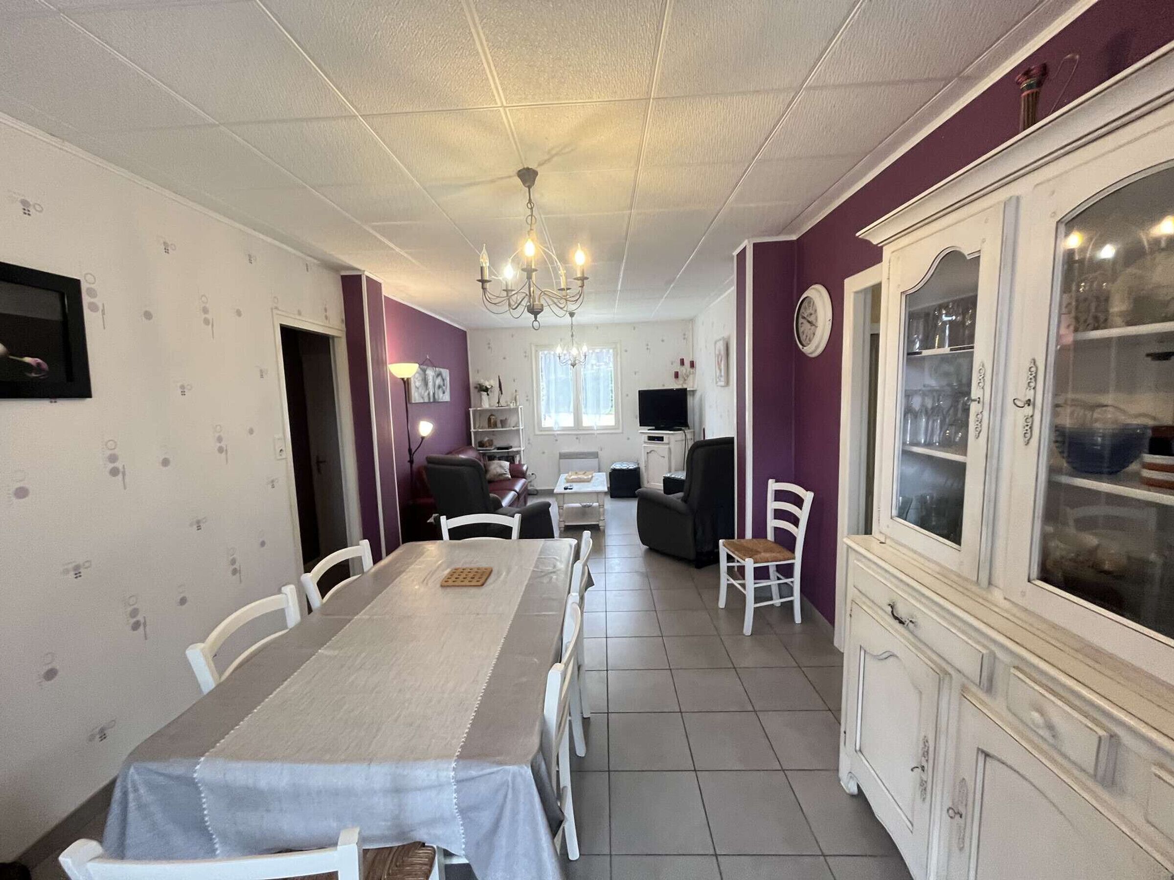 A dining area with a long table, chairs, a chandelier, a television, and a cabinet with glass doors.