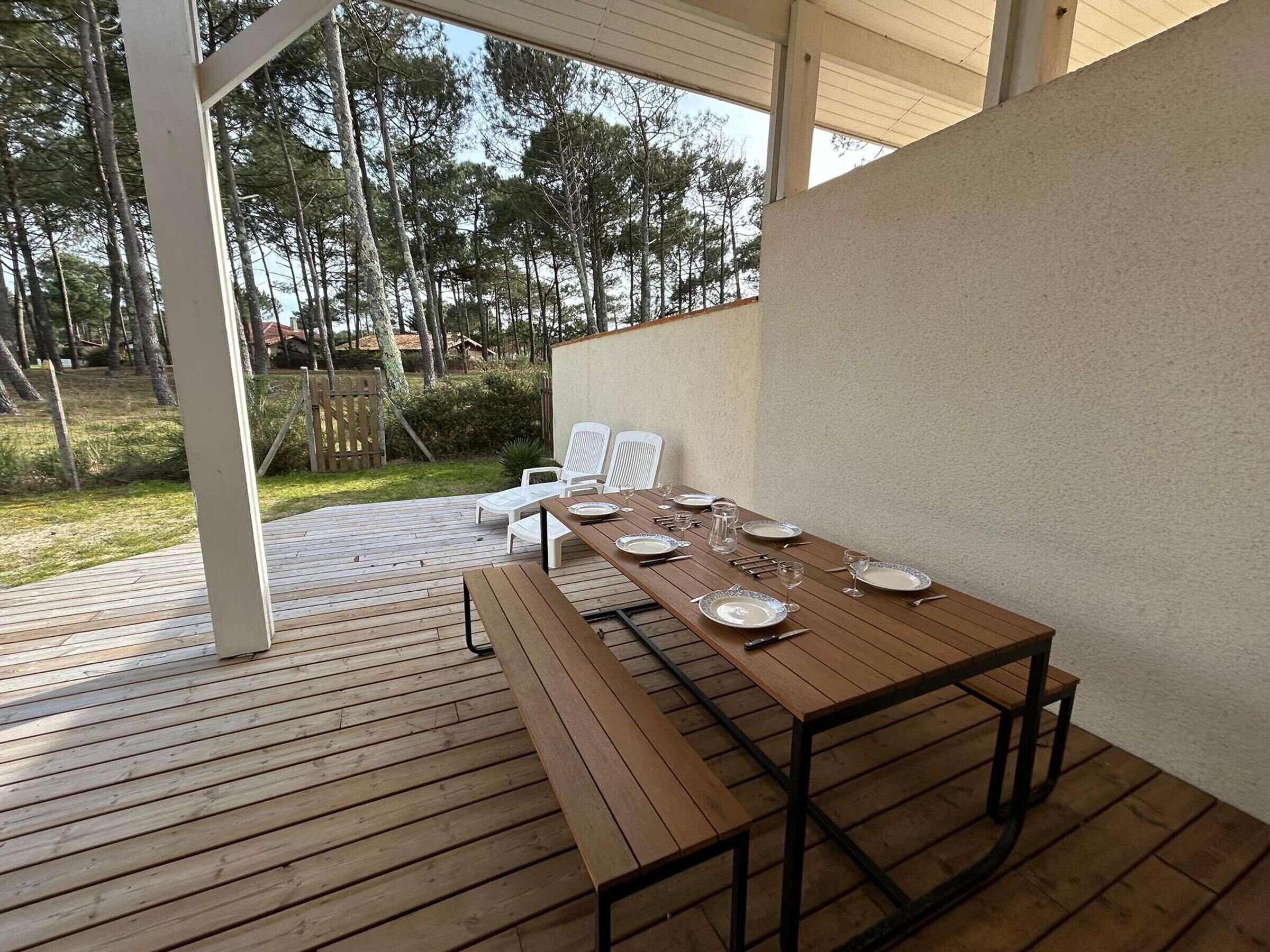 Une terrasse extérieure en bois, avec une table dressée pour le repas, entourée de chaises blanches, et une vue sur les arbres en arrière-plan.