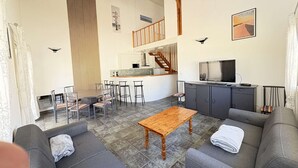 Amplia sala de estar con mesa de comedor, sofás grises, una mesa de centro de madera, cocina equipada con microondas y televisor.