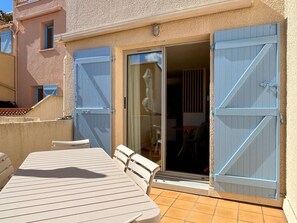 Un balcón con mesa y sillas, una puerta corredera de vidrio y un edificio con fachada texturizada.