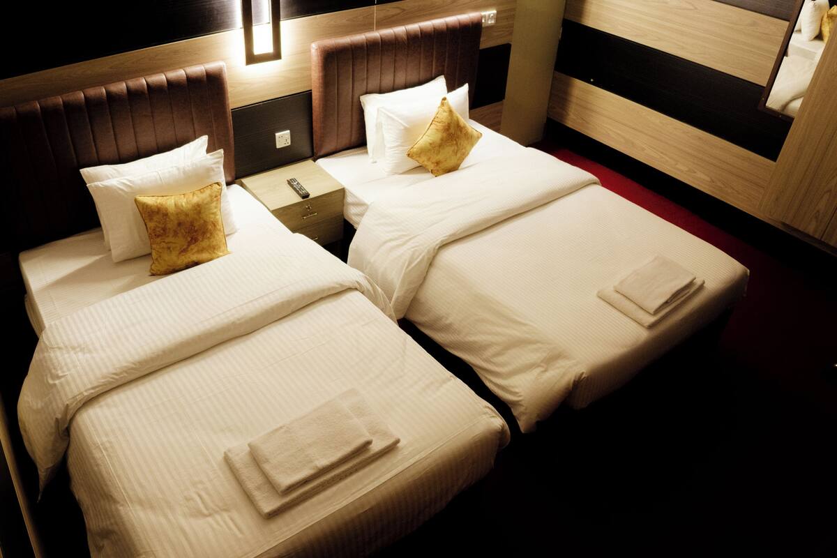 Una habitación de hotel con dos camas, cada una con sábanas y almohadas blancas, un cabecero de madera, una mesita de noche con un control remoto y una alfombra roja.