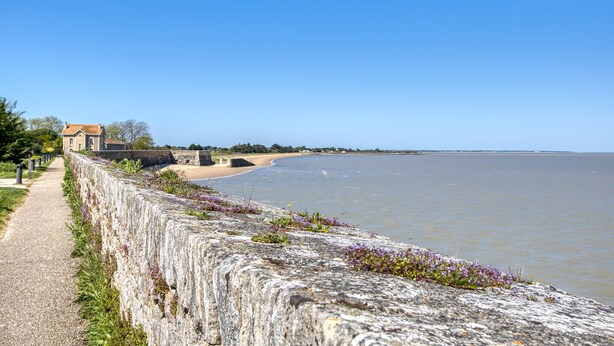 Une promenade en bord de mer bordée d’un muret en pierre, un bâtiment avec un toit rouge et une plage de sable.