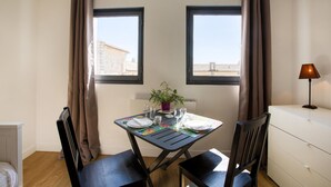 Un petit coin repas avec une table dressée pour deux personnes, deux fenêtres avec des rideaux, une plante en pot et une lampe sur une table de chevet.