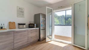Une cuisine moderne équipée d’un four, d’un micro-ondes et d’un réfrigérateur encastrés. On y trouve une planche à découper en bois et un plateau avec des bouteilles sur le comptoir. Une porte-fenêtre coulissante donne accès à un balcon avec vue sur les arbres.