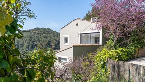 Une maison à deux étages dotée d’un balcon, entourée d’une végétation luxuriante et avec une colline boisée en arrière-plan.