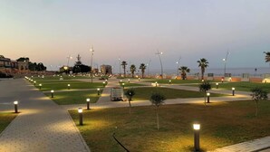 A well-lit pedestrian pathway with grass and trees, leading towards a body of water. - VFH The Castle Bay apartment and terrace (Santa Flavia)