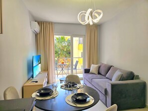Uma sala de estar moderna com sofá, mesa de jantar para dois, televisão e varanda com vista para palmeiras.