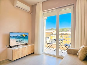 A modern living room with a flat-screen TV, wooden cabinet, and a balcony with a table and chairs.