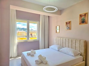 A bedroom with a large window, a bed with pillows, rolled towels, a ceiling light, and two framed pictures on the wall.