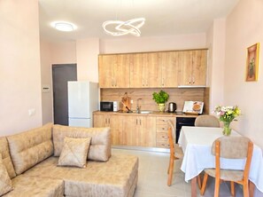 Una cocina moderna con armarios de madera, un refrigerador blanco, un microondas y un lavamanos. Hay un sofá beige con cojines, una mesa de comedor con sillas y un jarrón con flores sobre la mesa.