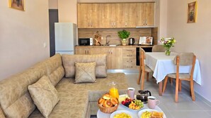 A modern living room with a sofa, a dining table with chairs, a kitchen area with wooden cabinets, and a hanging light fixture. - Apartment by the Sea in Golem 095 (Durres)