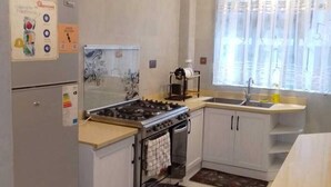 Et moderne køkken med gaskomfur, ovn og køleskab. Der er et vindue med lette gardiner og en vask med armatur.
