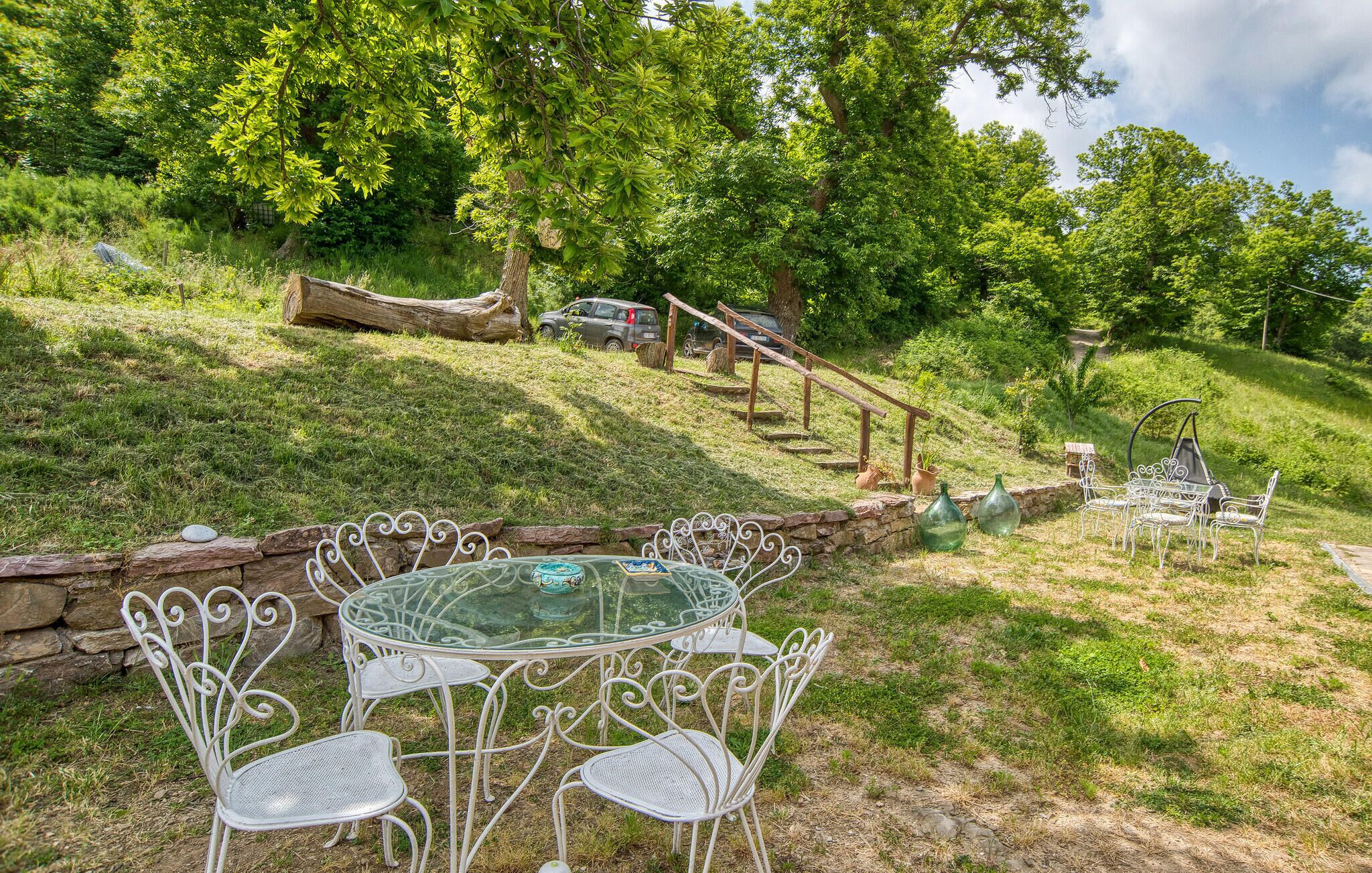 A rustic outdoor seating area with a glass-top table and white metal chairs, surrounded by a stone wall and lush greenery.