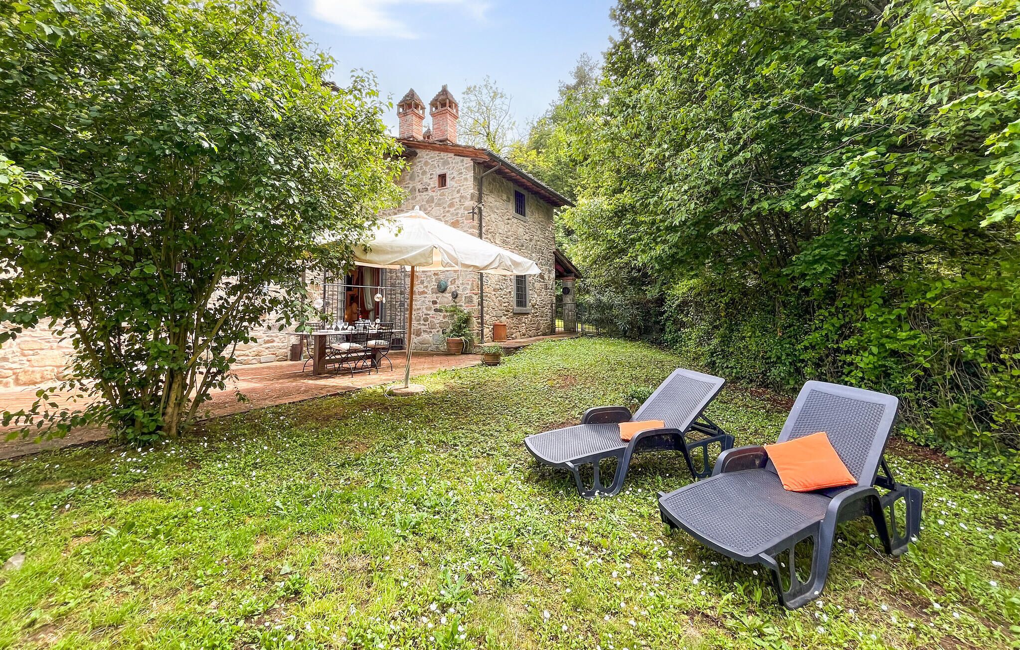 A stone building with a chimney, surrounded by lush greenery. There are two outdoor seating areas with umbrellas and chairs.