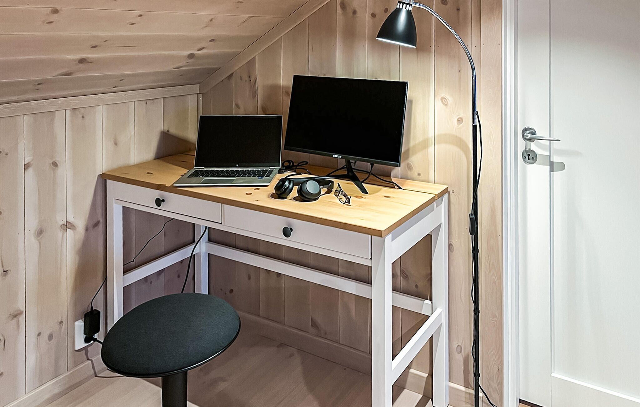 Ein Zimmer mit Holzvertäfelung, ausgestattet mit einem Schreibtisch, einem Laptop, einem Desktop-Monitor, Kopfhörern und einer Lampe.