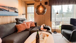 L’intérieur d’un chalet en bois chaleureux et accueillant, avec un canapé, une table basse sur laquelle se trouvent une bouteille et des verres, et une vue sur l’extérieur.