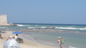 A beach with a castle-like structure, umbrellas, and people relaxing. - Romantic Trullo for 2  sea view pool,peaceful garden rustic  authentic Puglia (Carovigno)