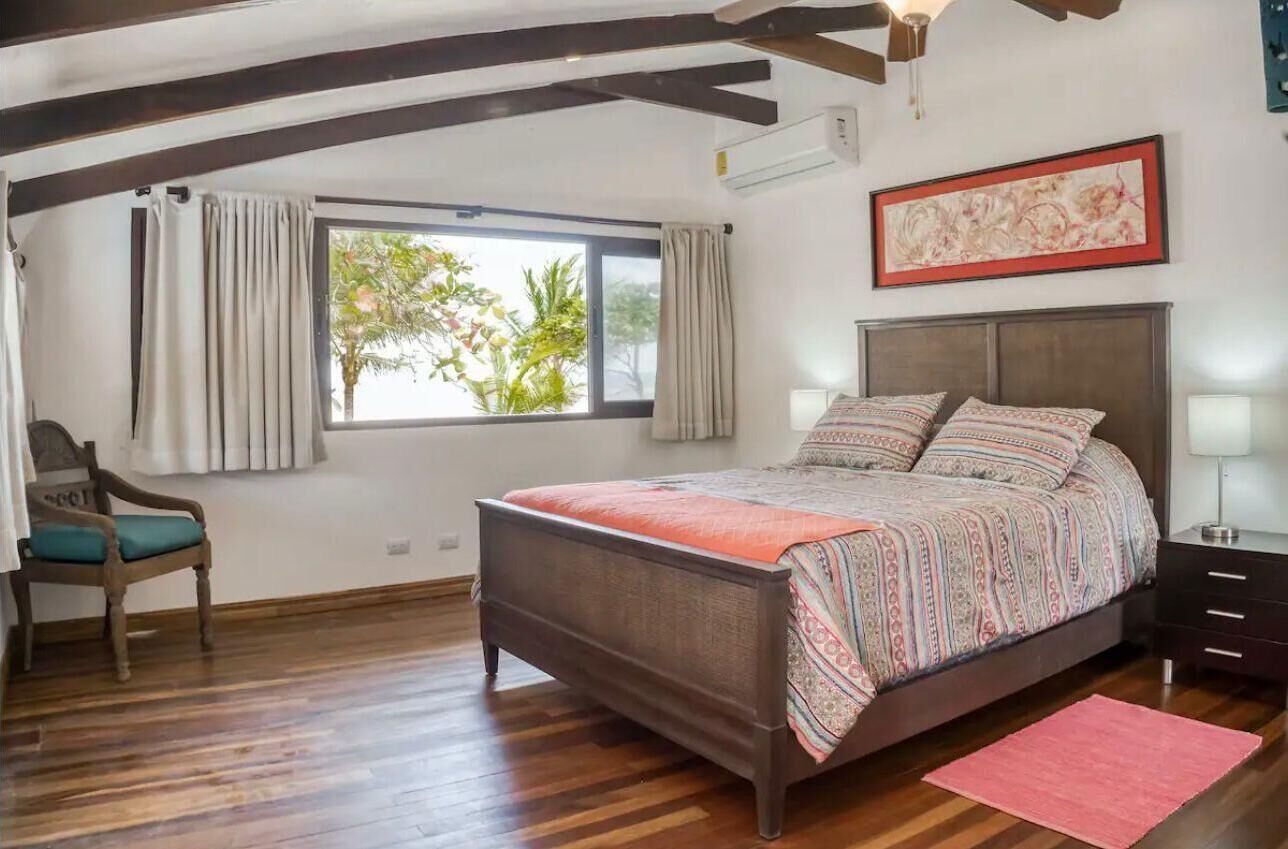 Une chambre à coucher avec un lit, une chaise, une fenêtre avec des rideaux, une table de chevet et un tableau encadré au mur.