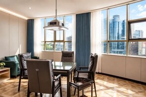 Habitación de hotel moderna con mesa de comedor, sillas, sofá y ventanas grandes que ofrecen vistas a la ciudad.