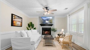 A living room with a white sofa, a fireplace, a ceiling fan, and a large window with blinds.