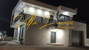 Una villa moderna con balcón, entrada bien iluminada y camino de entrada pavimentado.