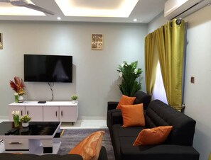 A modern living room with a flat-screen TV, a white entertainment unit, a black sofa with orange cushions, and a large green plant. - Spacious 3BR Apartment in Adentan With City Views (Adentan)