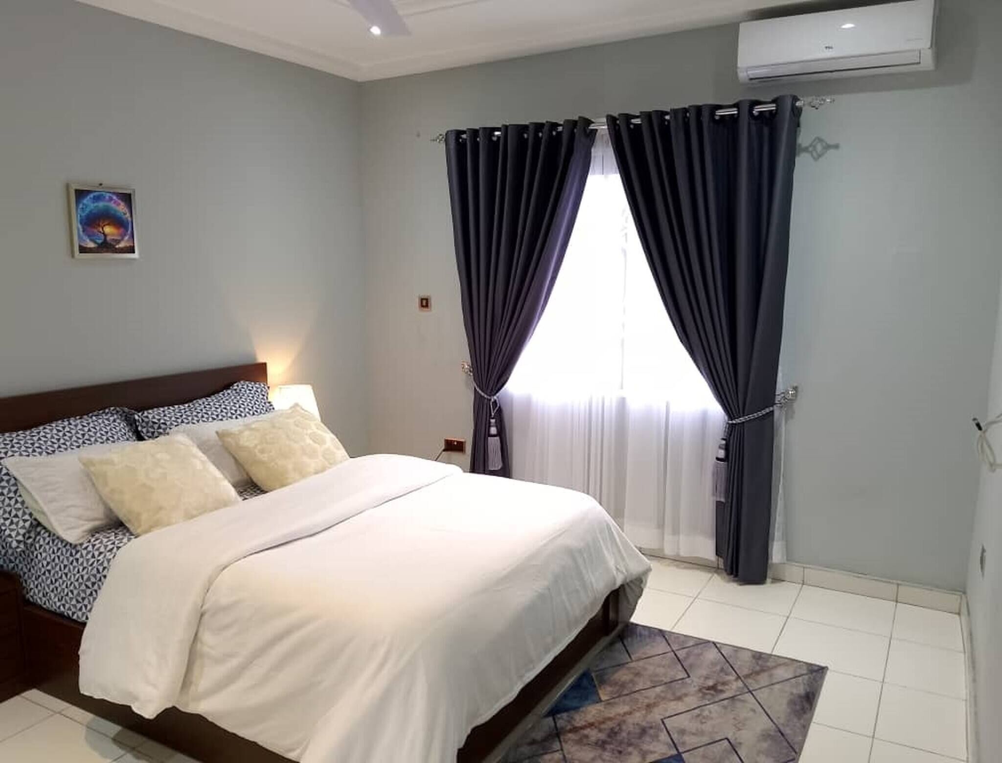 Un dormitorio con una cama, una mesita de noche, un aire acondicionado instalado en la pared y una ventana con cortinas.