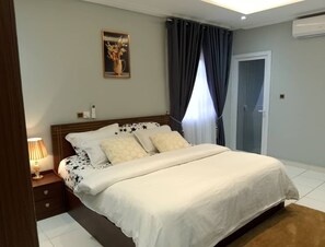 A bedroom with a large bed, a bedside table with a lamp, a framed picture on the wall, and a door with a glass panel. - Spacious 3BR Apartment in Adentan With City Views (Adentan)