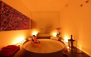 Een spa-ruimte met een bubbelbad, een muur met abstracte kunst, kaarsen en een tafel met een fles en een schaal vol aardbeien.