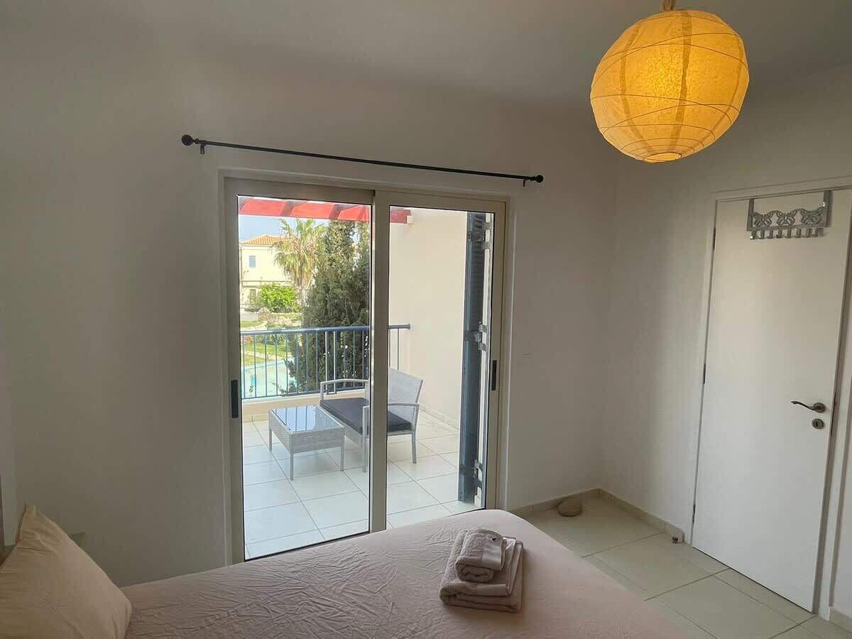 Une chambre avec un lit, une porte-fenêtre coulissante donnant sur un balcon équipé d’une table et de chaises, et un luminaire suspendu.
