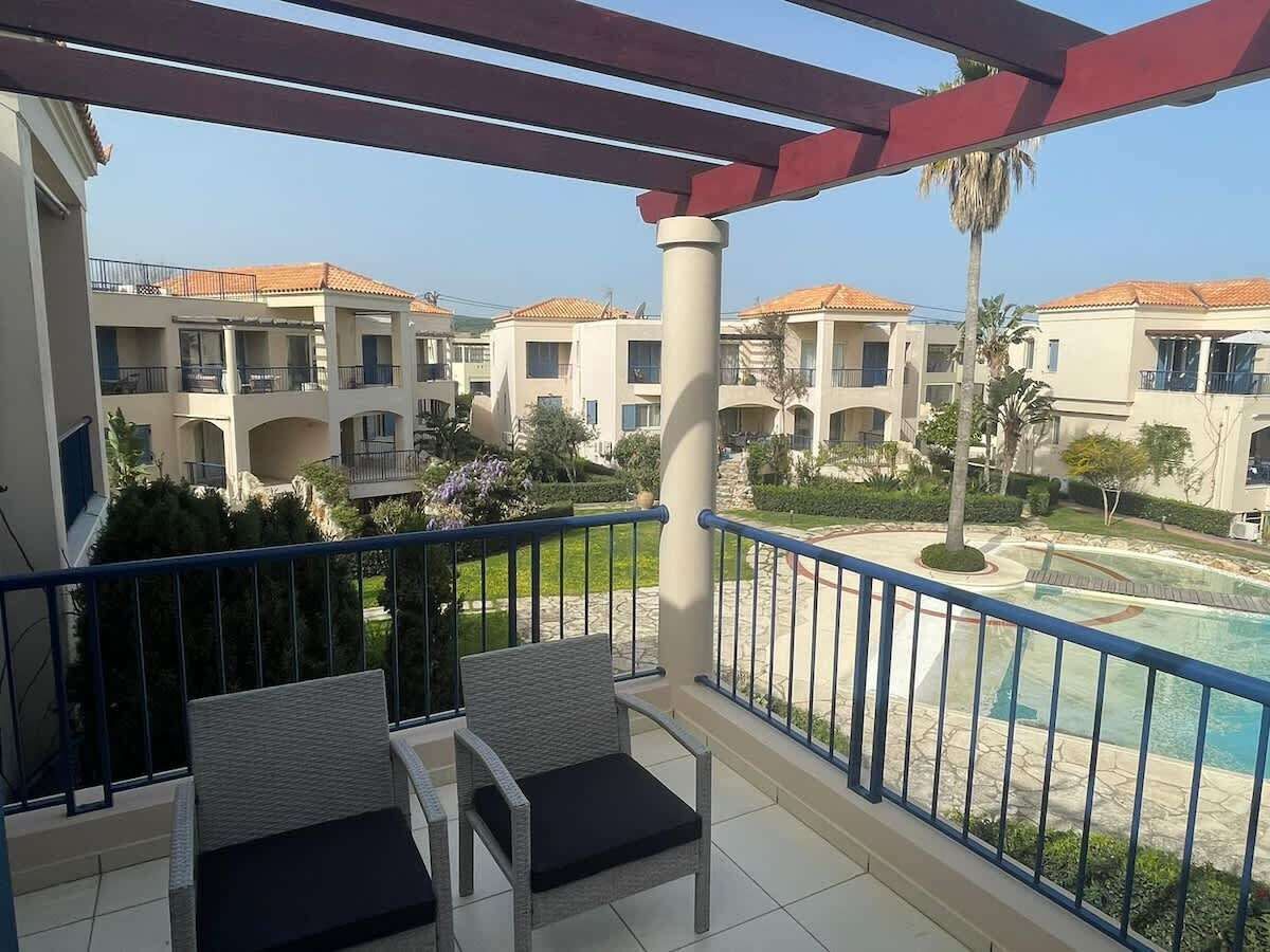A balcony with two wicker chairs, a pool, and a view of multi-story buildings with tiled roofs.