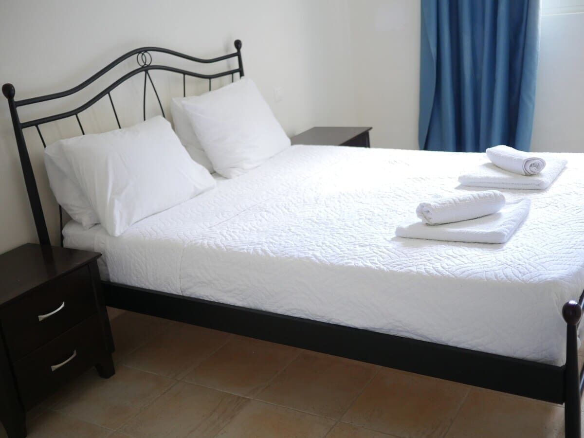 A neatly made bed with white linens and pillows, a dark wooden headboard, and a small bedside table with drawers.