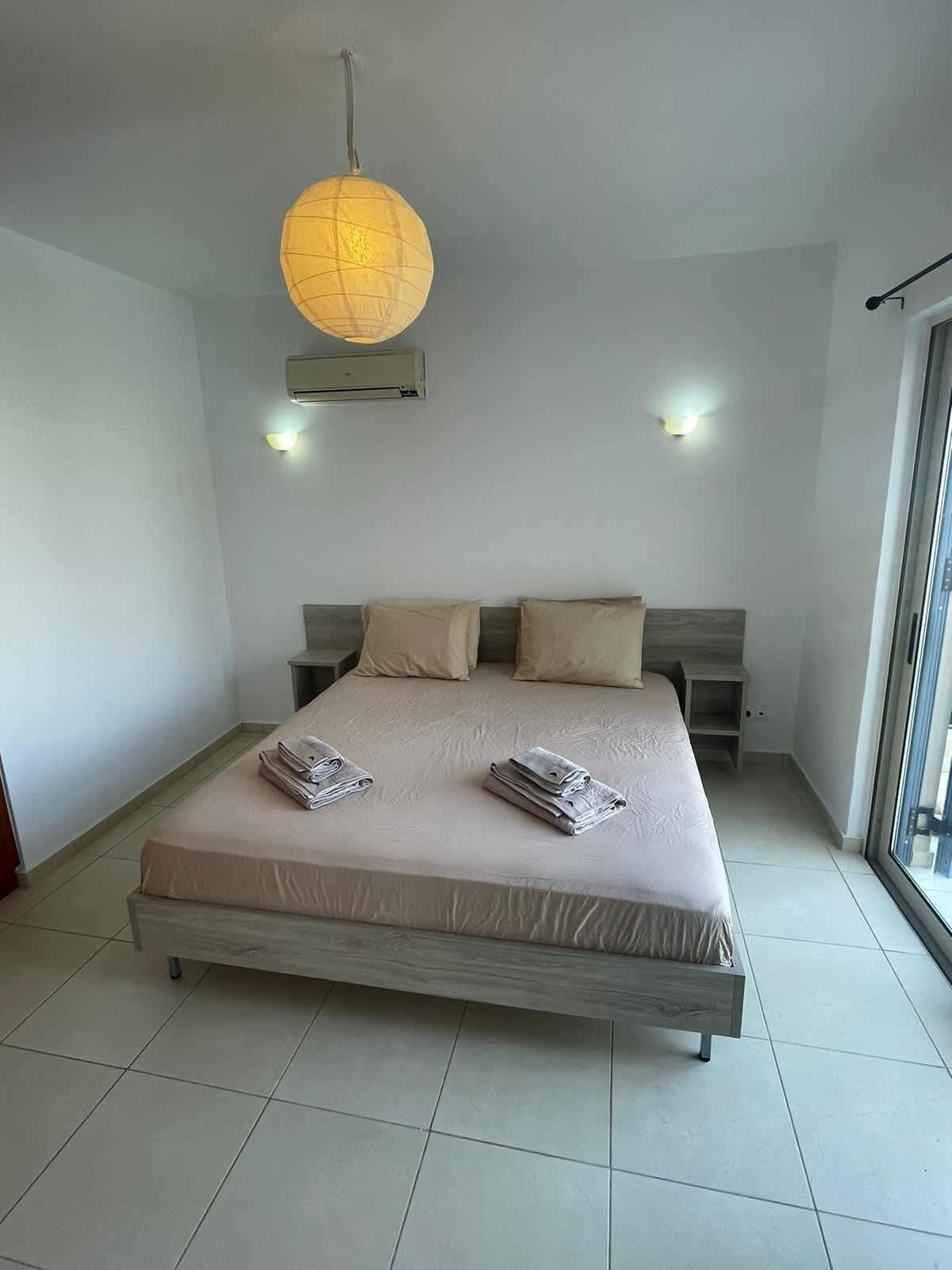 A modern bedroom with a large bed, two bedside tables, a hanging lamp, and a sliding glass door.