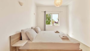 A minimalist bedroom with a wooden bed, a bedside table, a hanging light fixture, and a window with a view of greenery. - Cassiopeia 5 BR Villa on beach with pool (Pyrgos Psilonerou)