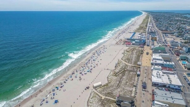 Vista aérea de una playa con una extensa franja de arena, algunos edificios y un cielo azul despejado.
