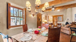 Un comedor con una mesa de madera dispuesta para cuatro personas, rodeada de sillas. Un candelabro cuelga sobre la mesa, y se ve una chimenea al fondo.