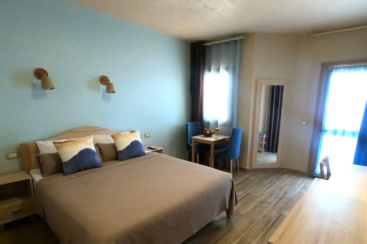 A hotel room with a bed, bedside tables, a desk, a chair, and a door leading to a balcony.