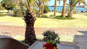 A wicker table with a vase of flowers, a chair, and a view of a beach with palm trees and the sea. - Mangrove Bay Resort (El Quseir)