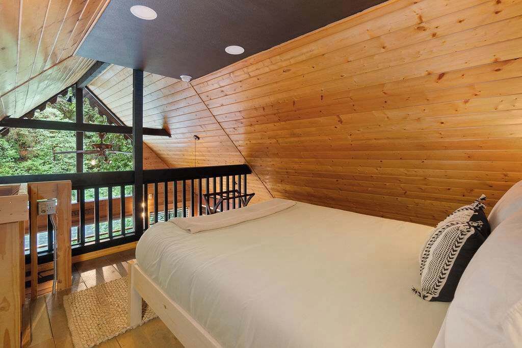 Un dormitorio con techo de madera, una cama con ropa blanca, una mesita pequeña y vistas a la vegetación exterior.