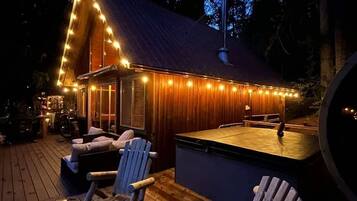 Una terraza de madera con sillas Adirondack, un jacuzzi y luces colgantes.