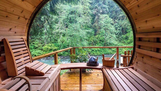 Eine Holzhütte mit rundem Fenster, das auf einen bewaldeten Bereich blickt. Im Inneren befinden sich Holzbänke und ein Tisch mit einem Eimer und einer Flasche.