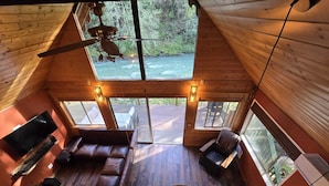 Eine Hütte mit Holzvertäfelung, Deckenventilator, Fernseher, Sofa, Esstisch und großen Fenstern mit Flussblick.