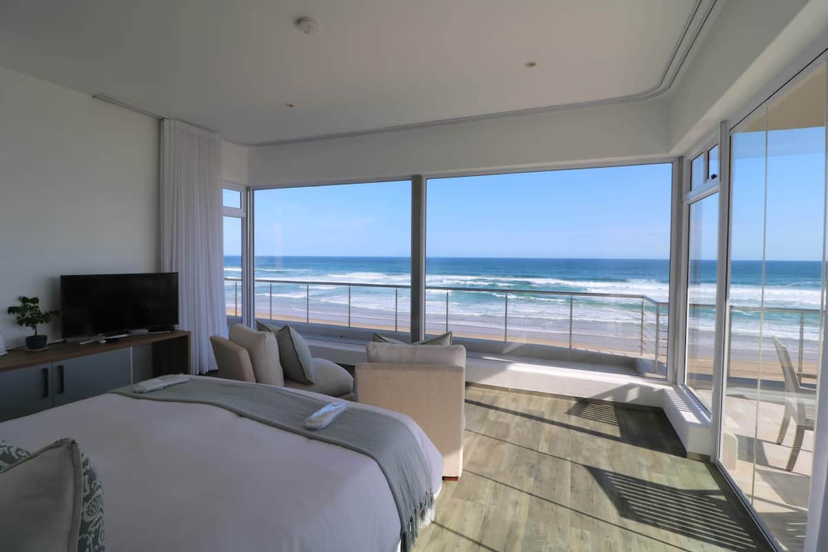 A modern hotel room with a large bed, a TV, and a balcony with a view of the beach and ocean.