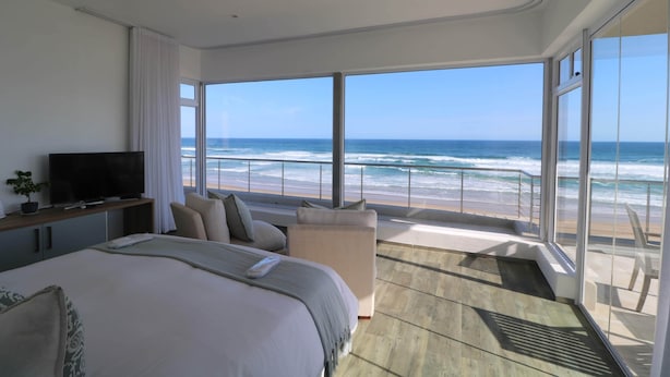 Ein modernes Hotelzimmer mit einem großen Bett, einem Fernseher und einem Balkon mit Blick auf Strand und Meer.