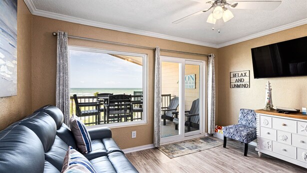 A living room with a leather sofa, a wooden coffee table, and a TV mounted on the wall. There is a sliding glass door leading to a balcony with beach views. - New Heated Pool & Hot tub! Adorable Condo! (Galveston)