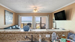 A beachside room with a view of the ocean, a ceiling fan, a flat-screen TV, and a 'Welcome to the Beach' sign. - New Heated Pool & Hot tub! Adorable Condo! (Galveston)