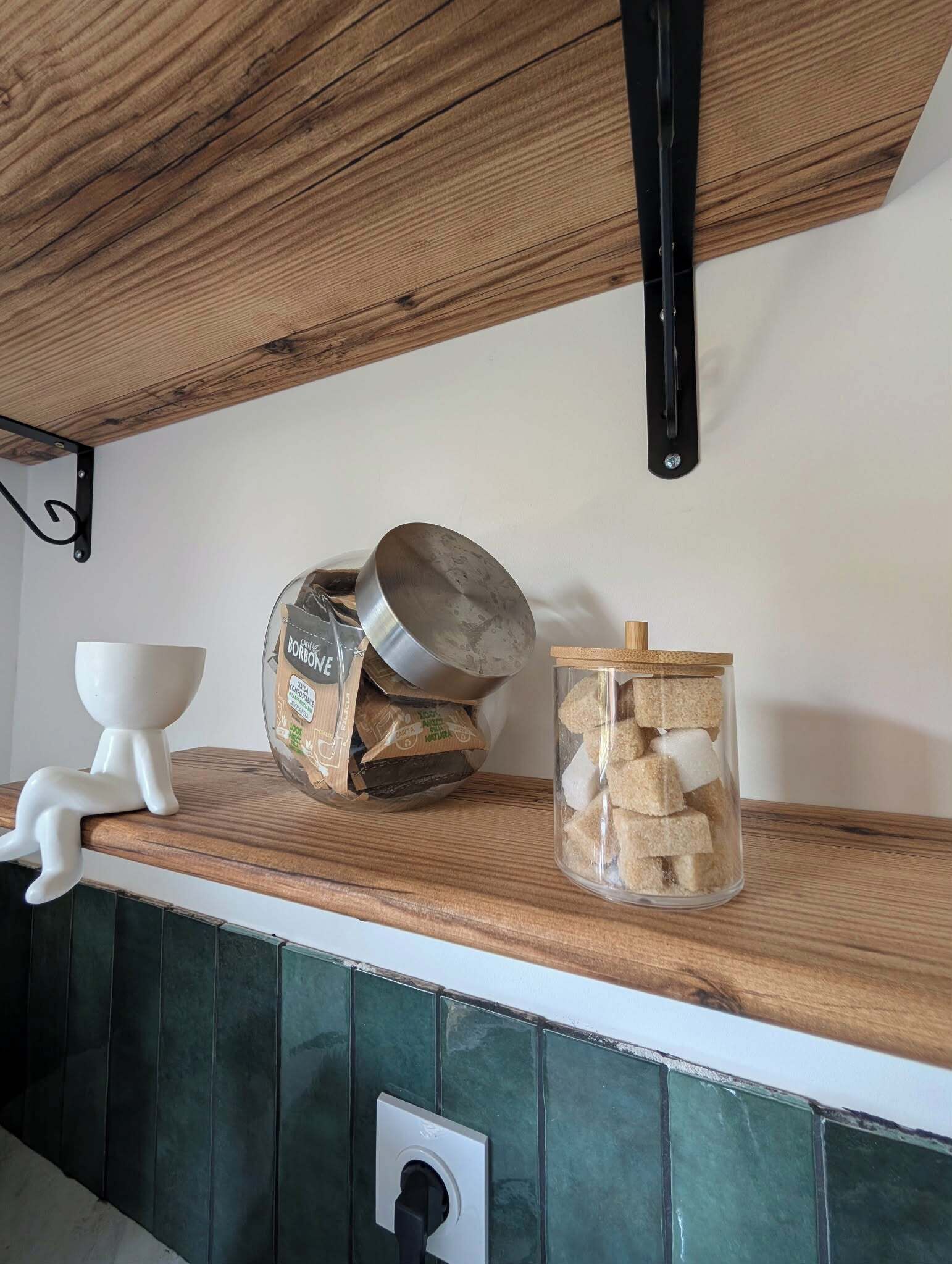 Un comptoir de cuisine avec une étagère en bois, une tasse en céramique blanche, un bocal en verre contenant des morceaux de sucre et une casserole en métal.
