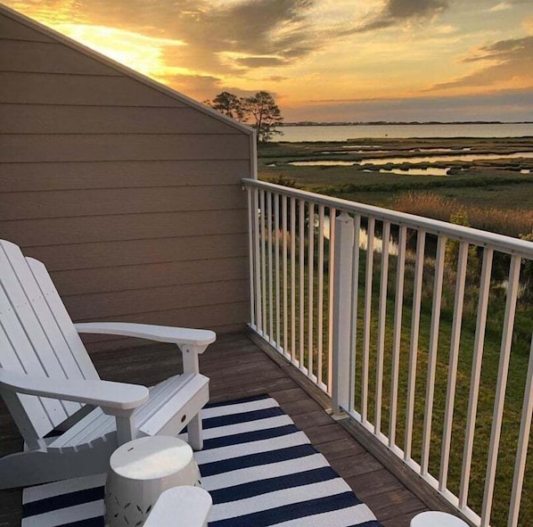 A balcony with white chairs and a striped rug, overlooking a marshy landscape with a sunset.