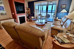 A living room with a sofa, a coffee table, and a fireplace. There is a television on the wall and a view of the outdoors through large windows.