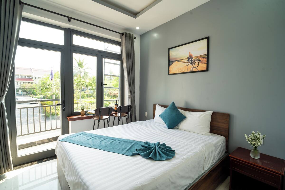 A hotel room with a large bed, a view of the outdoors, a painting on the wall, and a small table with a vase.